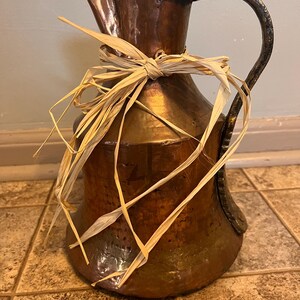 May include: A hammered copper pitcher with a decorative handle and a raffia bow. The pitcher has a dark brown patina and a unique shape, suitable for rustic or farmhouse decor.