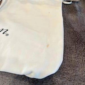 May include: A white drawstring bag with a small brown stain. The bag is made of a soft material and is partially visible, with a woven straw bag in the background.