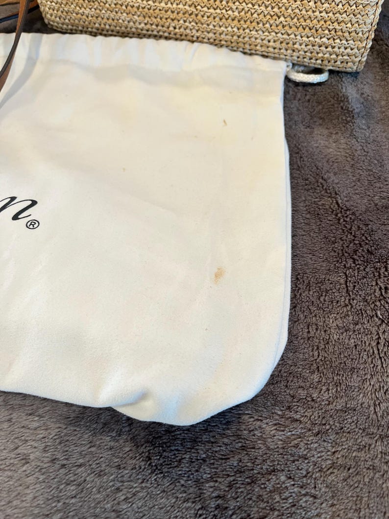 May include: A white drawstring bag with a small brown stain. The bag is made of a soft material and is partially visible, with a woven straw bag in the background.