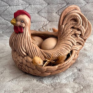 May include: A ceramic hen-shaped container in shades of tan, with a red comb and yellow beak. The container holds two eggs and decorative dried grasses. The hen's tail feathers are intricately detailed.