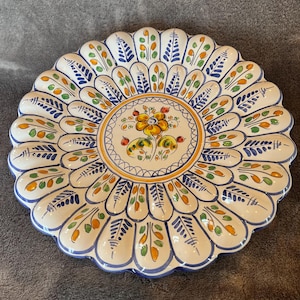 May include: A decorative ceramic plate with a scalloped edge. The plate features a central floral design in yellow, orange, and red, surrounded by a border of blue, green, and orange leaf-like patterns. The plate has a white background.