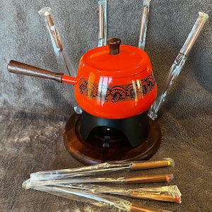 May include: An orange fondue pot with a wooden handle and lid knob, set on a wooden base. The pot has a black decorative pattern. Several fondue forks are included, wrapped in plastic.