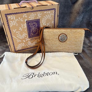 May include: A tan woven crossbody bag with a brown leather strap and a silver decorative emblem. The bag is accompanied by a matching box and a white dust bag, all bearing the Brighton brand name.