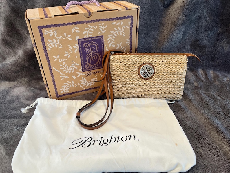 May include: A tan woven crossbody bag with a brown leather strap and a silver decorative emblem. The bag is accompanied by a matching box and a white dust bag, all bearing the Brighton brand name.