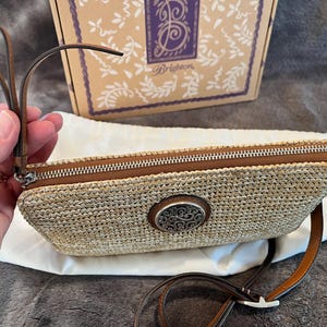 May include: A woven straw clutch with a brown leather zipper and strap. The clutch features a decorative silver-toned oval emblem. A brown box with a logo is in the background.