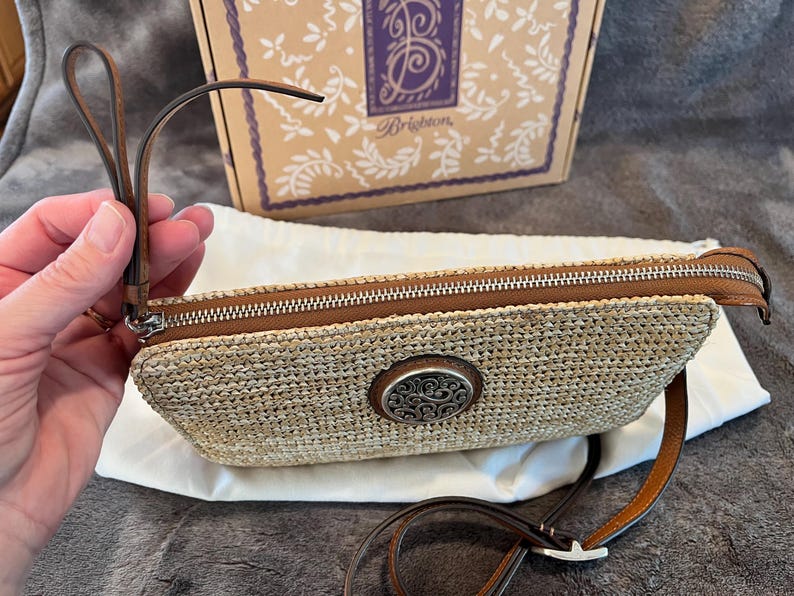 May include: A woven straw clutch with a brown leather zipper and strap. The clutch features a decorative silver-toned oval emblem. A brown box with a logo is in the background.