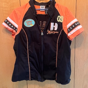 May include: Black and orange zip-up motorcycle jacket with short sleeves. The jacket features the text "Cruiser Girls" and "Harley Davidson Motorcycles". The sleeves have white stripes with black stars. The number "03" is embroidered on the right side.