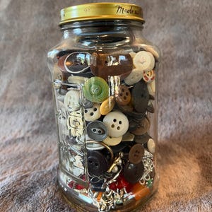 May include: A clear glass Mason jar filled with a variety of assorted buttons in different colors, shapes, and sizes. The jar features a gold-colored metal lid with the text Made with printed on the top edge and embossed glass lettering.