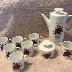 May include: A white porcelain tea set featuring a red rose design with teal and gray accents. The set includes a coffee pot, sugar bowl, creamer, and eight mugs. The tea set is displayed on a pink textured surface.