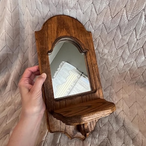 May include: A wall-mounted wooden mirror featuring a medium brown stained finish with a prominent wood grain. The mirror has an arched top and includes a small, integrated horizontal shelf extending from the base, supported by a decorative bracket.