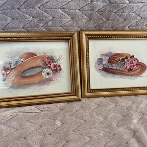 May include: Two framed watercolor paintings of hats. One hat is a light orange color with pink and white flowers. The other hat is brown with a blue and white patterned ribbon and pink flowers. Both paintings are in gold frames.
