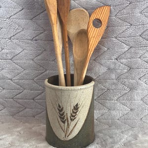 May include: A cylindrical ceramic utensil crock featuring a two-tone beige and dark green glaze with a brown wheat stalk design on the front. The crock is filled with five assorted wooden kitchen spoons and spatulas of varying shapes and sizes.