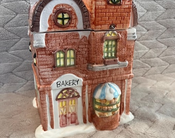 Op de afbeelding: Een keramische koekjespot in de vorm van een bakkerijgebouw met meerdere verdiepingen, rode bakstenen muren, een witte boogdeur, een blauw-wit gestreepte luifel boven een etalage en een dak met witte sneeuwaccenten en het woord BAKERY.