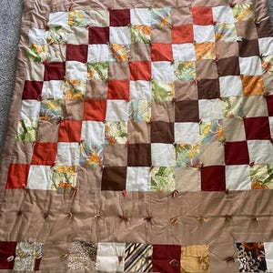 May include: A patchwork quilt with a grid pattern of squares in various colors, including brown, red, orange, and floral prints. The quilt is made of fabric and has a tan border with decorative knots.
