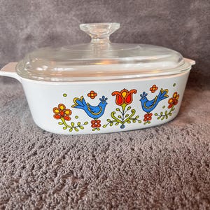 May include: A white rectangular casserole dish with a clear glass lid and integrated side handles. The exterior features a colorful pattern of two blue birds, orange flowers, and green stems printed along the center of the white ceramic body.