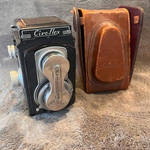 May include: A black Ciro-flex twin-lens reflex camera with silver metal dials and a lens cover, accompanied by a brown leather carrying case. The camera body features the text Ciro-flex and Made in U.S.A. printed on the top front panel.