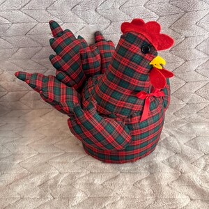 May include: A stuffed fabric chicken ornament featuring a dark green and red plaid pattern. The chicken has a bright red felt comb and wattle, a yellow beak, a small black button eye, and a thin red ribbon tied in a bow around its neck.