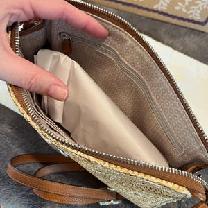 May include: A straw-colored clutch with a brown leather trim and strap. The interior features a patterned lining and a zippered pocket. The clutch is open, revealing a package inside. The zipper is silver.