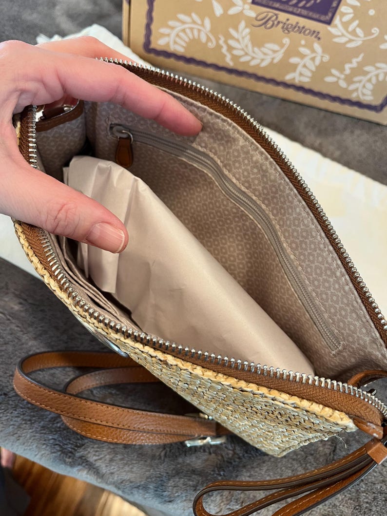 May include: A straw-colored clutch with a brown leather trim and strap. The interior features a patterned lining and a zippered pocket. The clutch is open, revealing a package inside. The zipper is silver.