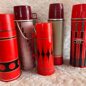 May include: Five vintage vacuum flasks in various designs. One is bright red with black circular patterns, another has a pink tiled motif, a third is orange with black diamond shapes, one is beige with a red base, and one is red with black stripes.