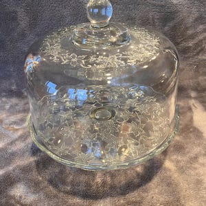 May include: A clear glass cake stand with a domed lid. The lid has a round knob on top and is decorated with a floral pattern. The base is a round plate with a raised edge, also decorated with the same floral pattern.