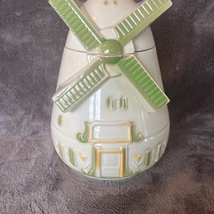 May include: A ceramic windmill-shaped cookie jar. The jar is primarily off-white with green and gold accents. The blades of the windmill are green, and the base features a door and windows. The jar is designed to resemble a traditional Dutch windmill.
