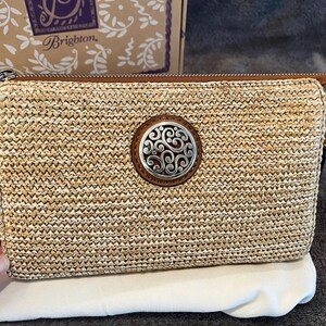 May include: A rectangular woven straw clutch with a brown leather zipper pull and a decorative silver and brown circular emblem. The clutch is on a white cloth, with a Brighton box in the background.