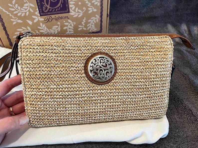 May include: A rectangular woven straw clutch with a brown leather zipper pull and a decorative silver and brown circular emblem. The clutch is on a white cloth, with a Brighton box in the background.