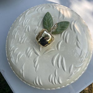May include: A round, cream-colored ceramic lid featuring a raised, embossed leaf pattern across its surface. The center is adorned with a metallic gold-toned apple-shaped handle, accented by two small, textured green ceramic leaves at its base.
