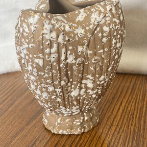 May include: A decorative ceramic vase with a unique shape and textured surface. The vase is primarily tan with white and gold accents. The vase has a pedestal base and a heart-shaped opening at the top.