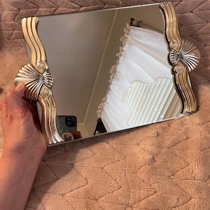 May include: A rectangular vanity mirror featuring a reflective glass surface framed by two ornate metallic side panels. Each side panel displays a prominent shell motif at the base, accented by vertical, flowing, wave-like ridges in a polished gold tone.