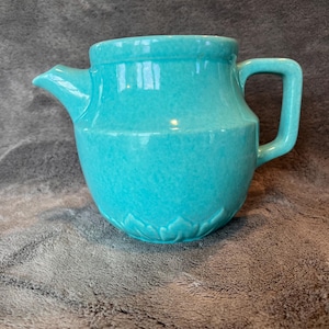 May include: A turquoise ceramic teapot with a glossy, speckled finish. The vessel features a wide, rounded body, a short spout, and a sturdy handle. The base of the teapot is decorated with a subtle, embossed petal or leaf pattern encircling the bottom.