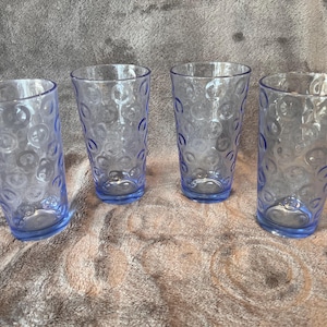 May include: Four clear, blue-tinted drinking glasses. Each glass has a cylindrical shape with a slightly flared rim and a blue base. The glass surface is decorated with a pattern of small, raised circles.