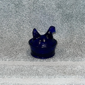 May include: A cobalt blue glass hen on a nest figurine. The hen is perched atop a round, textured base. The figurine is made of a dark blue glass and is likely a decorative item.