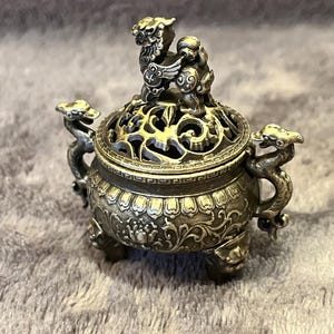 May include: A silver-colored incense burner with intricate detailing. The burner features a decorative lid with a sculpted animal figure on top. The sides are adorned with dragon-shaped handles and ornate patterns, resting on three small feet.