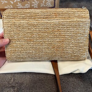 May include: A rectangular woven straw clutch with a brown leather zipper pull and a matching brown leather strap. The clutch is resting on a white cloth.