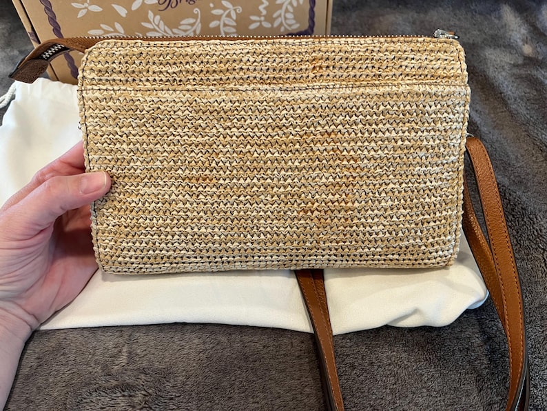 May include: A rectangular woven straw clutch with a brown leather zipper pull and a matching brown leather strap. The clutch is resting on a white cloth.