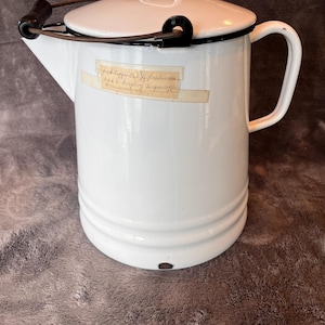 May include: A white enamel coffee pot with a cylindrical body, a side handle, and a matching lid. It features a dark metal swing handle with a black wooden grip, a spout, and a rectangular adhesive label with handwritten text on the front surface.