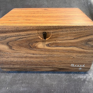 May include: A rectangular wooden bread box with a natural brown wood grain pattern. The front panel features a small black latch in the center and the word Bread written in white script lettering above a small white starburst icon on the bottom right.