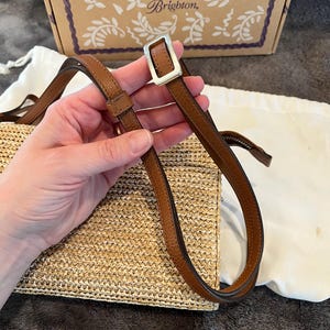 May include: A woven straw handbag with a brown leather strap and silver buckle. The bag is rectangular and has a light tan color. The strap is held by a hand. The background includes a box with the word "Brighton" on it.