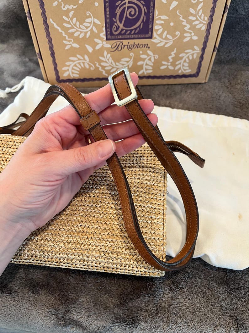 May include: A woven straw handbag with a brown leather strap and silver buckle. The bag is rectangular and has a light tan color. The strap is held by a hand. The background includes a box with the word "Brighton" on it.