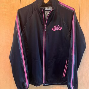 May include: Black zip-up jacket with pink stripes down the sleeves and sides. The jacket features a pink logo on the chest and a patterned lining. The jacket is hanging on a blue hanger.