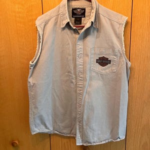 May include: A sleeveless, button-down shirt in a light tan color, hanging on a red hanger. The shirt features a Harley-Davidson logo patch on the chest pocket and a similar logo on the collar. The edges of the sleeves are frayed.