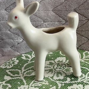 May include: A cream-colored ceramic deer figurine with red eyes. The deer has a small opening on its back, suggesting it could be used as a planter or decorative vase. The figurine is standing on a green and white patterned surface.