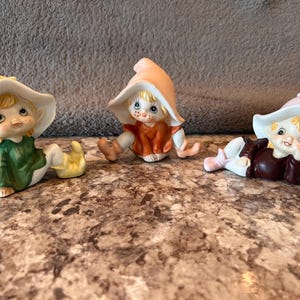 May include: Three ceramic elf figurines with pointed hats and blonde hair. The first elf wears a green outfit, the second an orange outfit, and the third a dark brown outfit. Each elf has large blue eyes, rosy cheeks, and a distinct pose.