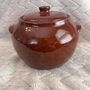 May include: A round, dark brown ceramic pot with a glossy finish, featuring two small side handles and a matching removable lid with a central knob. The pot has a wide, bulbous body and a smooth, reflective surface that catches light.
