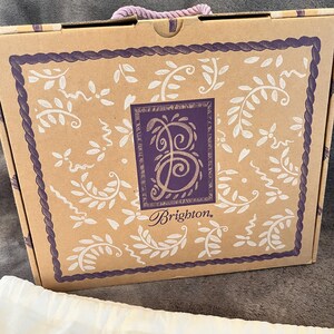 May include: A brown cardboard box with a purple rope handle. The box has a decorative border and a central design featuring the word "Brighton" and a stylized letter "B". White floral patterns adorn the box.