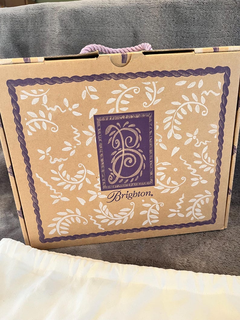 May include: A brown cardboard box with a purple rope handle. The box has a decorative border and a central design featuring the word "Brighton" and a stylized letter "B". White floral patterns adorn the box.