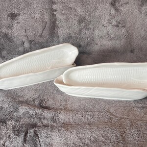 May include: Two white ceramic corn-shaped serving dishes. The dishes are detailed with a textured surface to resemble corn kernels. The dishes are placed on a gray surface.