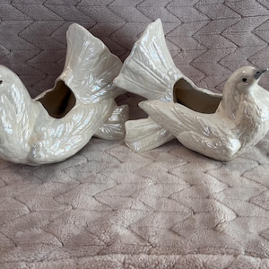 May include: Two white ceramic bird-shaped planters with an iridescent, pearlescent finish. Each planter features detailed molded feathers, a prominent tail, and a hollow opening on the back for holding small items or plants, presented as a pair.